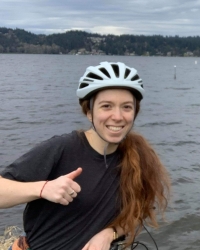 Picture of a woman giving a thumbs up in front of a lake. She is wearing a bike helmet and has long red hair.